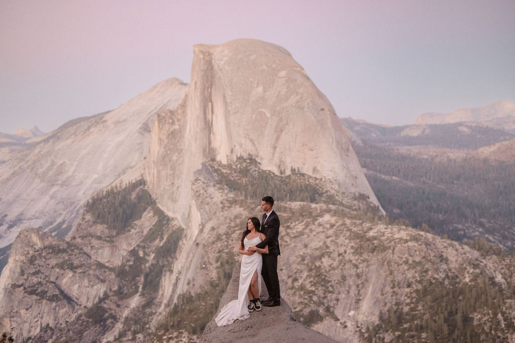 A couple in formal attire stands on a rocky ledge with a scenic mountain backdrop at sunset.