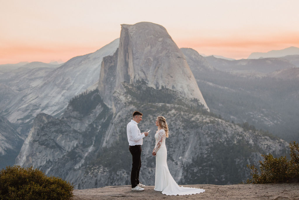 A couple, dressed in a wedding gown and suit, stands on a mountain ledge, with a vast mountain range and Half Dome in the backdrop during sunset. what to pack for a yosemite elopement