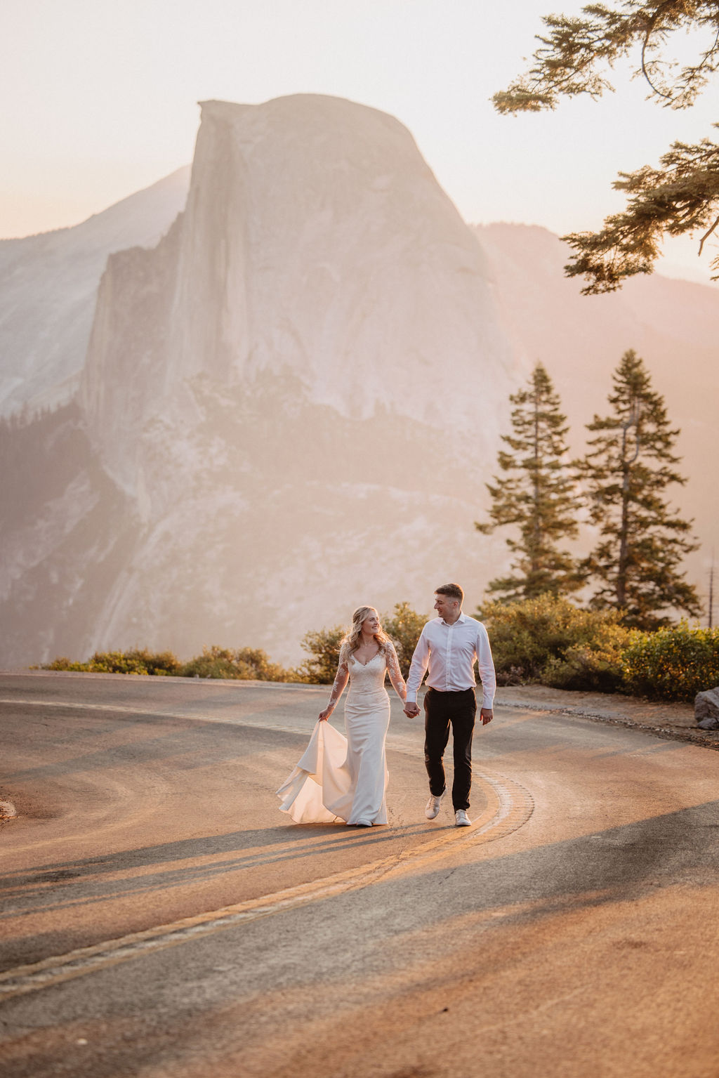 A couple in wedding attire holding hands and walking on a curved mountain road with a large rock formation and trees in the background. 