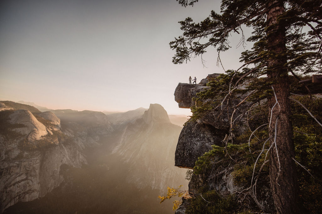 Two people stand on a cliff edge overlooking a vast, mountainous landscape at sunset, with a tree in the foreground.