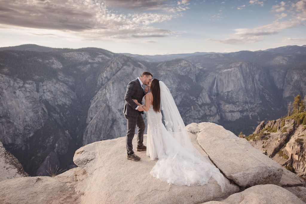 A bride and groom in wedding attire share a kiss on a rocky cliff with a mountainous landscape in the background.