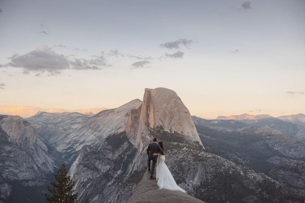 A bride and groom in wedding attire share a kiss on a rocky cliff with a mountainous landscape in the background.