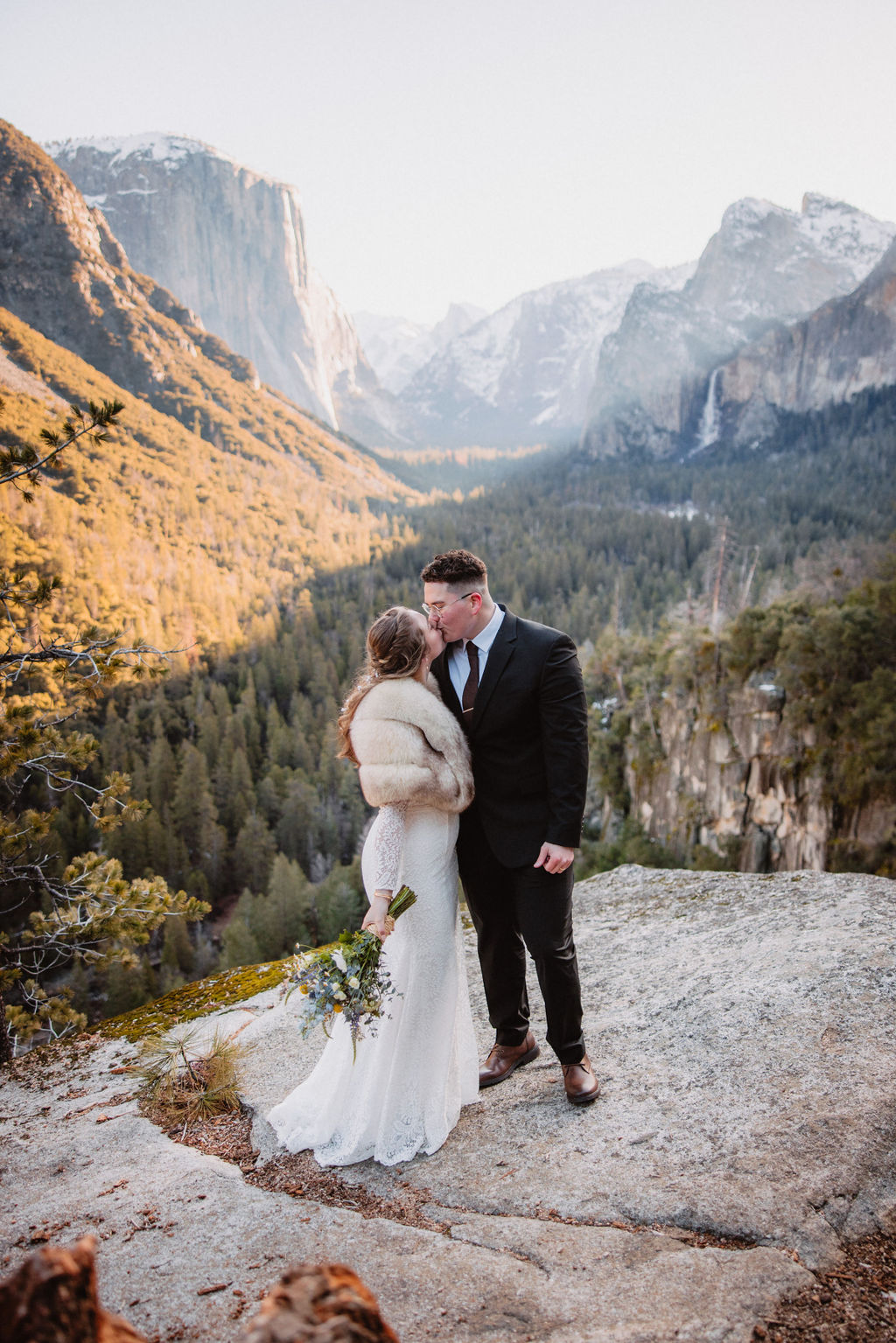 A couple in wedding attire share an embrace outdoors on a sunlit mountain path with snowy surroundings and forested hills in the background for their yosemite elopement