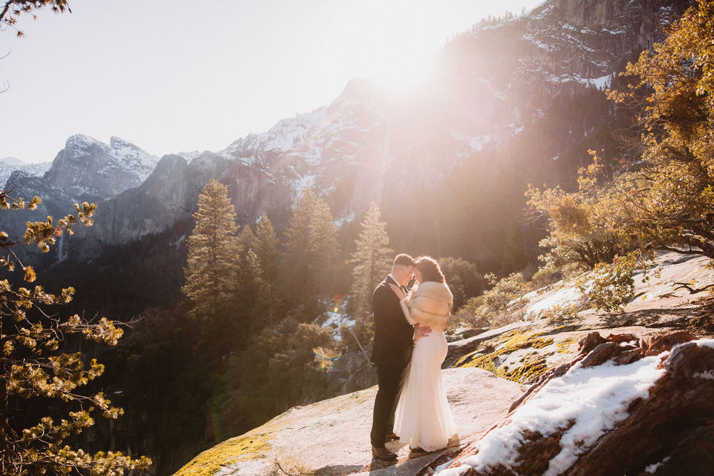 A couple in wedding attire share an embrace outdoors on a sunlit mountain path with snowy surroundings and forested hills in the background for their yosemite elopement 