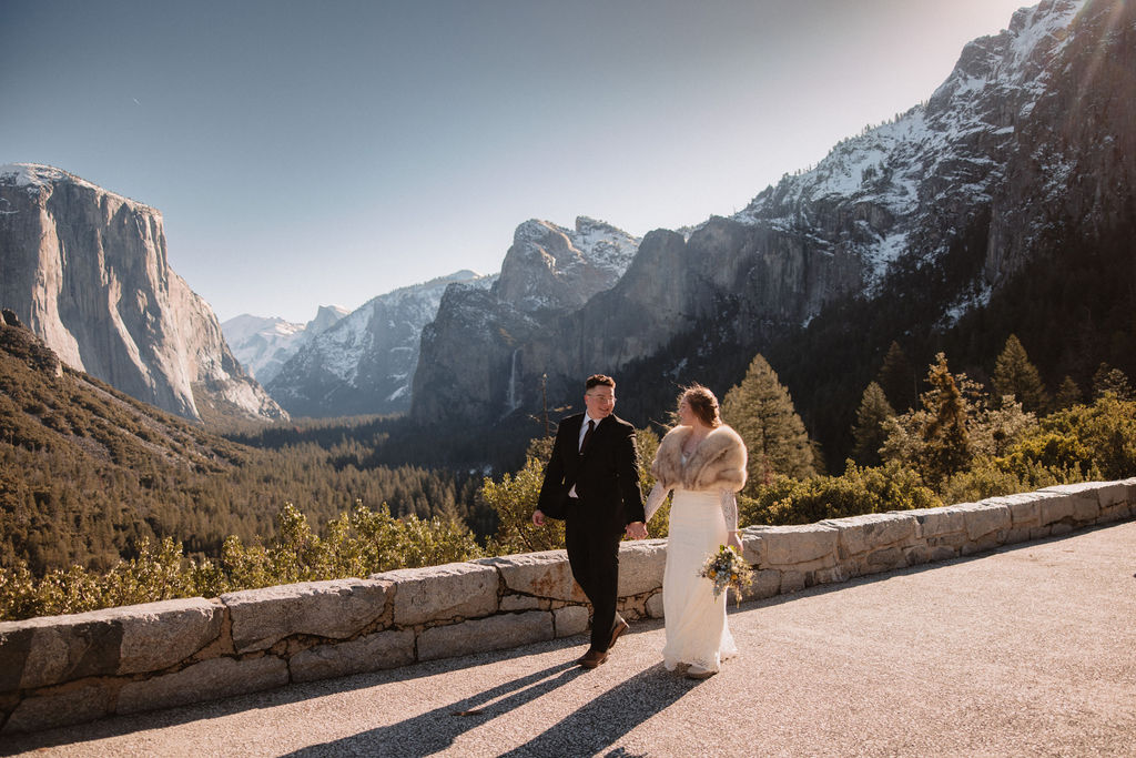 A couple in wedding attire share an embrace outdoors on a sunlit mountain path with snowy surroundings and forested hills in the background for their yosemite elopement 