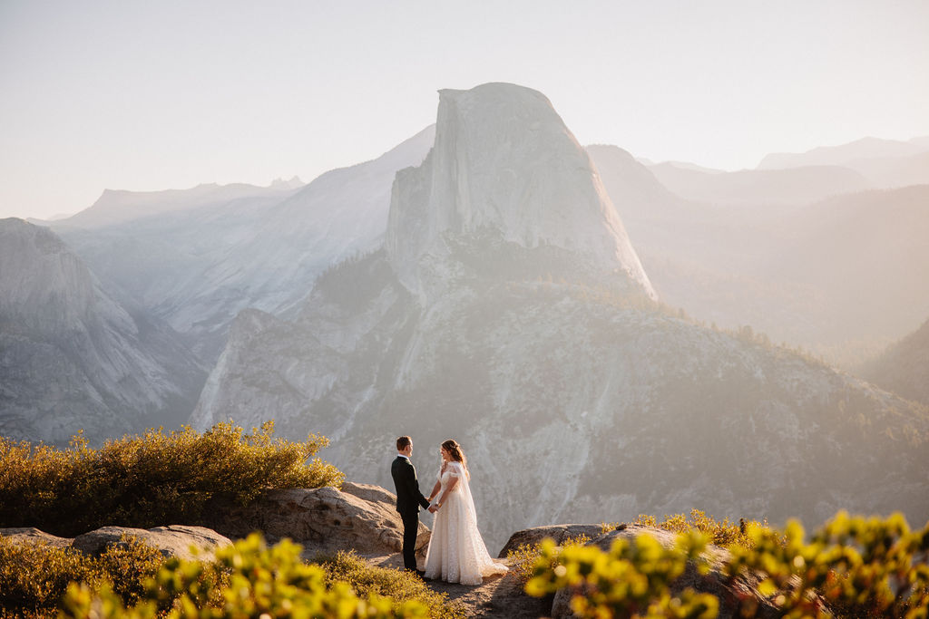 A couple in wedding attire stand together on a rocky overlook with a mountain landscape in the background at Yosemite national park at glacier point and mariposa grove 