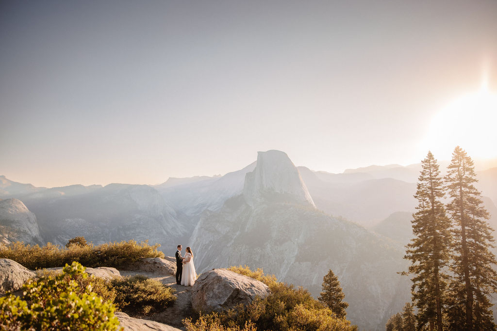 A couple in wedding attire stand together on a rocky overlook with a mountain landscape in the background at Yosemite national park at glacier point and mariposa grove 