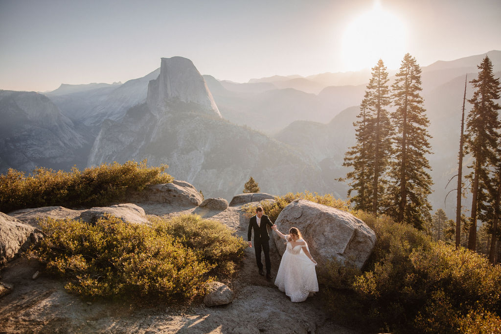 A couple in wedding attire stand together on a rocky overlook with a mountain landscape in the background at Yosemite national park at glacier point and mariposa grove 