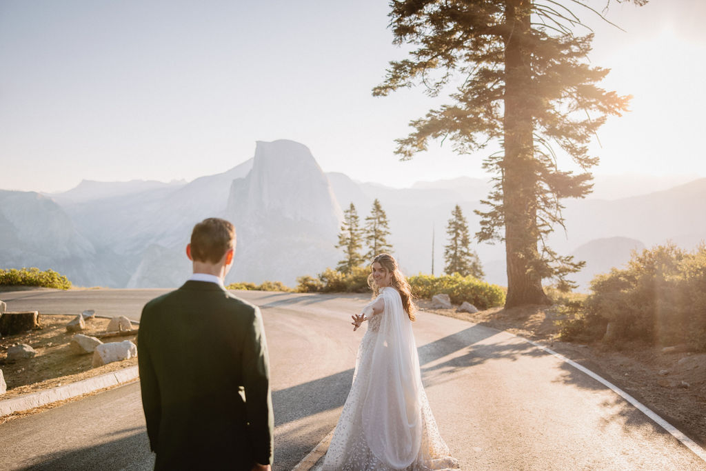 A couple in wedding attire stand together on a rocky overlook with a mountain landscape in the background at Yosemite national park at glacier point and mariposa grove 