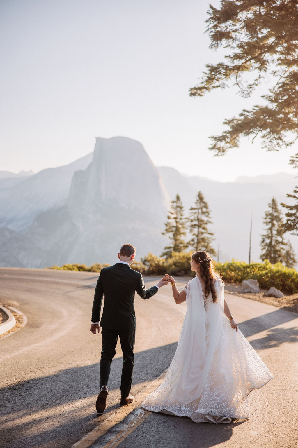 A couple in wedding attire stand together on a rocky overlook with a mountain landscape in the background at Yosemite national park at glacier point and mariposa grove 