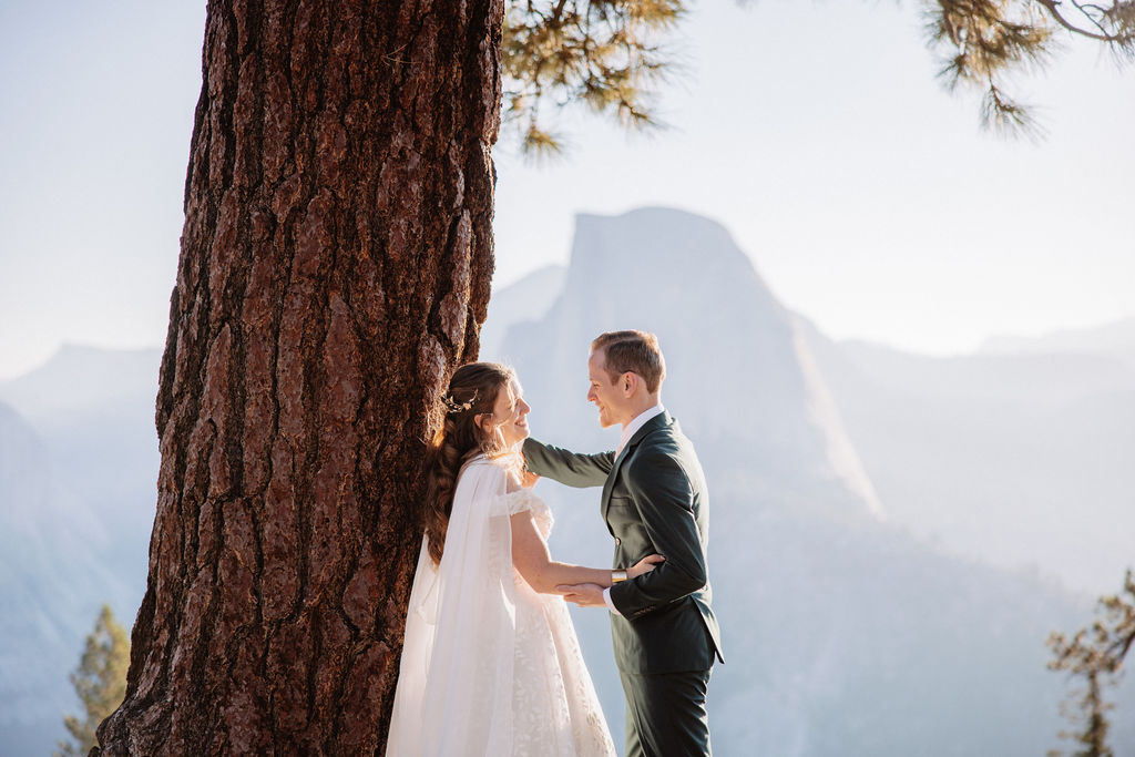 A couple in wedding attire stand together on a rocky overlook with a mountain landscape in the background at Yosemite national park at glacier point and mariposa grove 