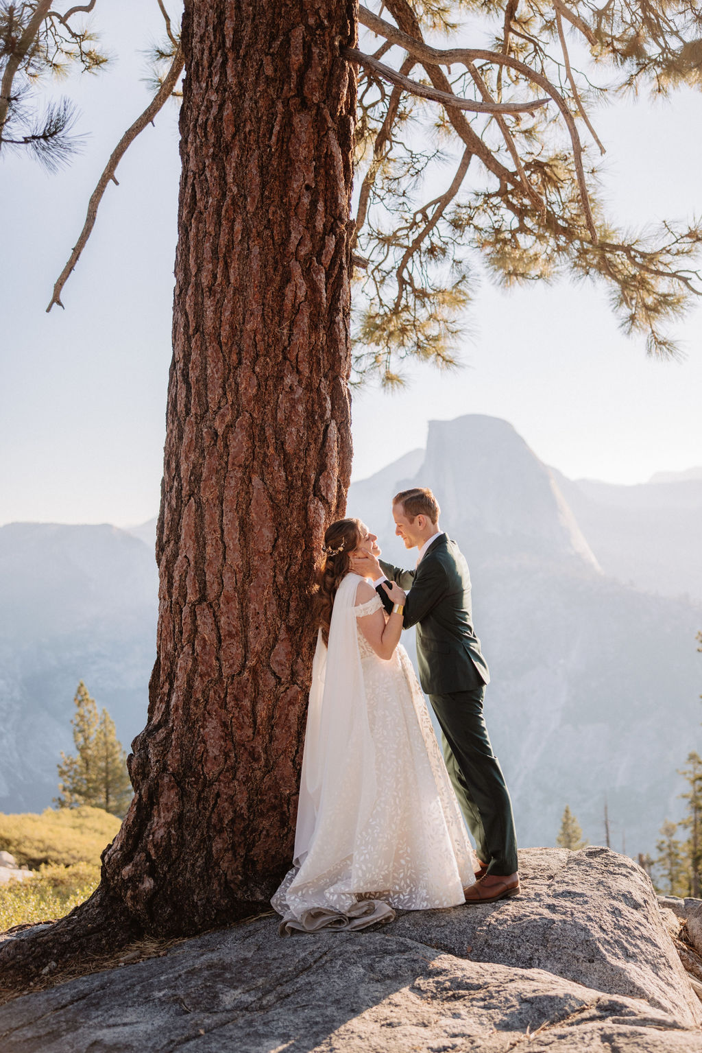 A couple in wedding attire stand together on a rocky overlook with a mountain landscape in the background at Yosemite national park at glacier point and mariposa grove 