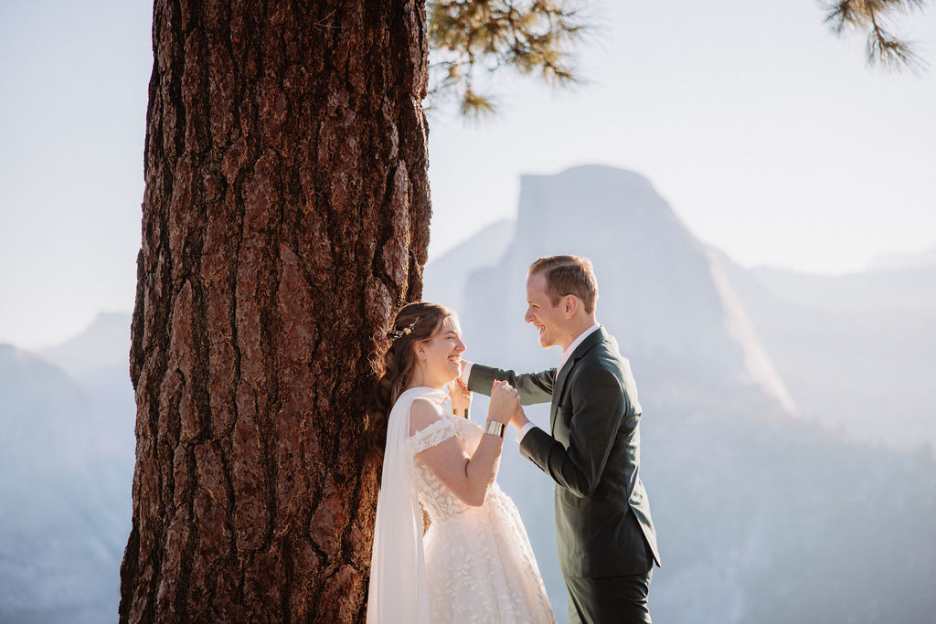 A couple in wedding attire stand together on a rocky overlook with a mountain landscape in the background at Yosemite national park at glacier point and mariposa grove 