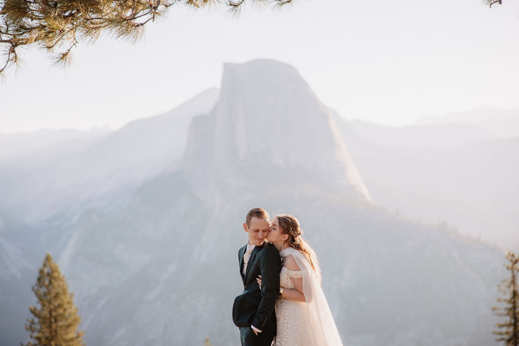 A couple in wedding attire stand together on a rocky overlook with a mountain landscape in the background at Yosemite national park at glacier point and mariposa grove 