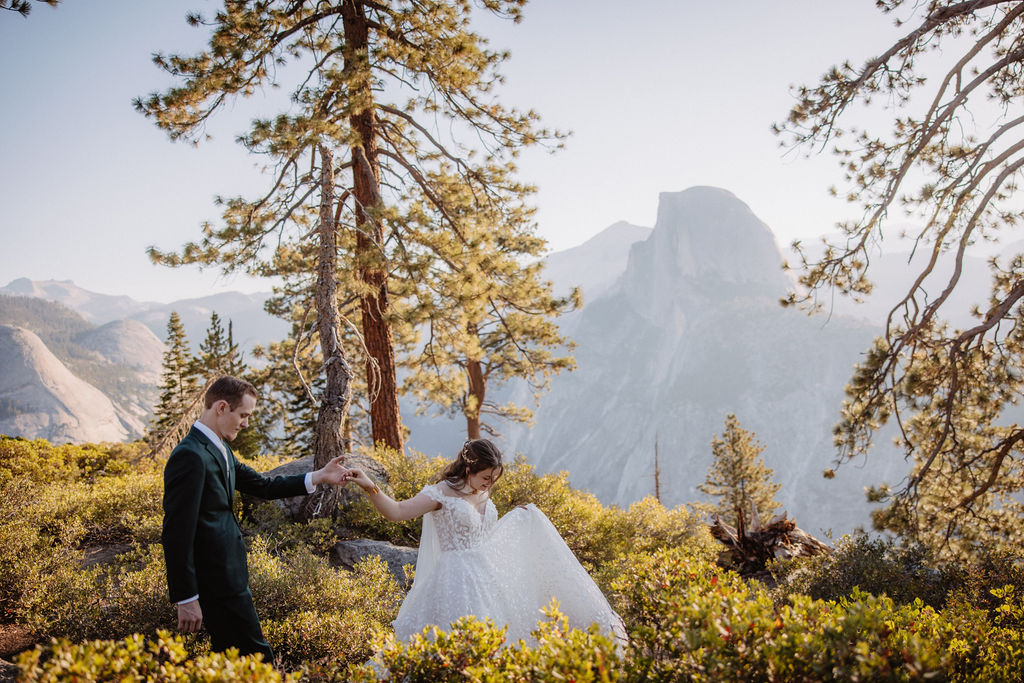 A couple in wedding attire stand together on a rocky overlook with a mountain landscape in the background at Yosemite national park at glacier point and mariposa grove