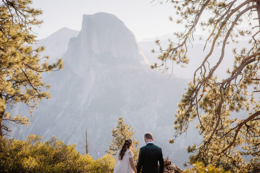 A couple in wedding attire stand together on a rocky overlook with a mountain landscape in the background at Yosemite national park at glacier point and mariposa grove