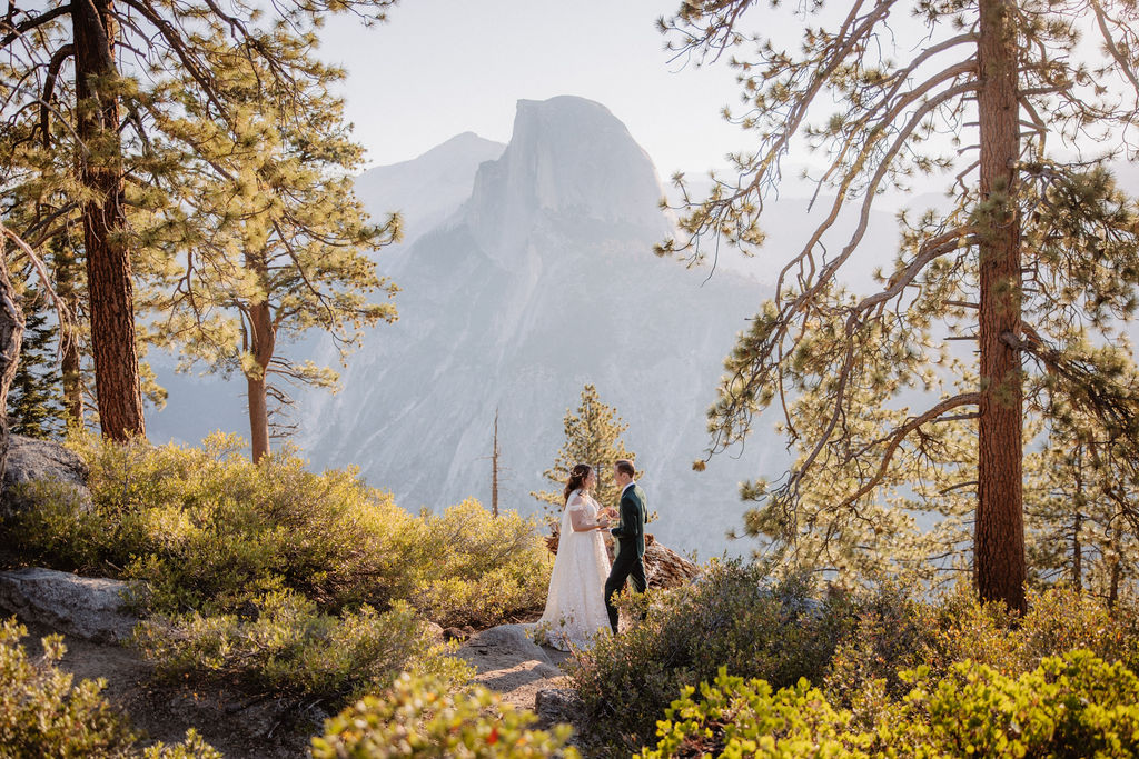 A couple in wedding attire stand together on a rocky overlook with a mountain landscape in the background at Yosemite national park at glacier point and mariposa grove