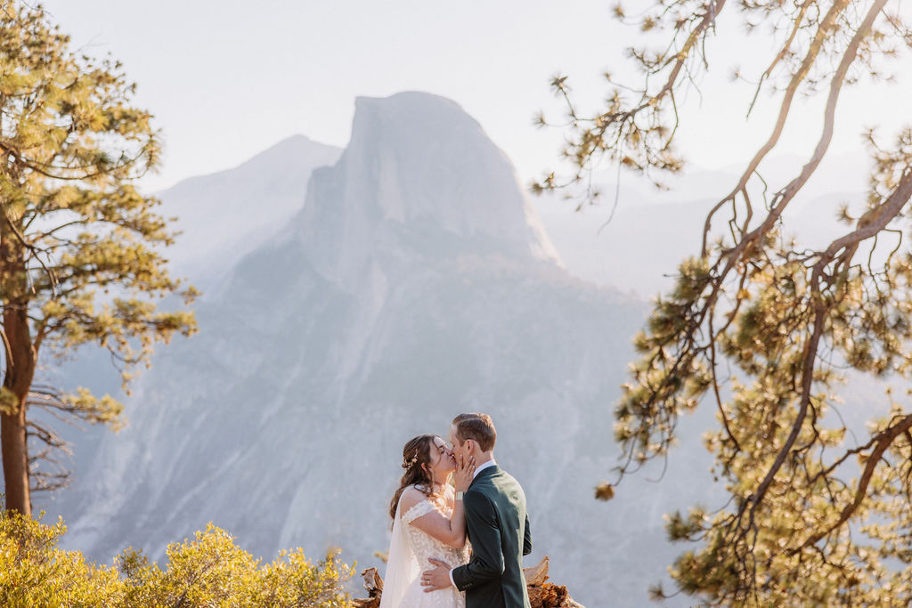 A couple in wedding attire stand together on a rocky overlook with a mountain landscape in the background at Yosemite national park at glacier point and mariposa grove