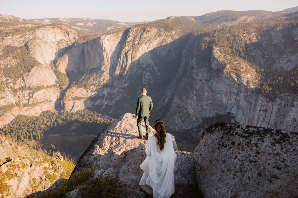 A couple in wedding attire stand together on a rocky overlook with a mountain landscape in the background at Yosemite national park at glacier point and mariposa grove