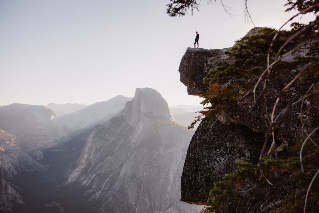 A couple in wedding attire stand together on a rocky overlook with a mountain landscape in the background at Yosemite national park at glacier point and mariposa grove