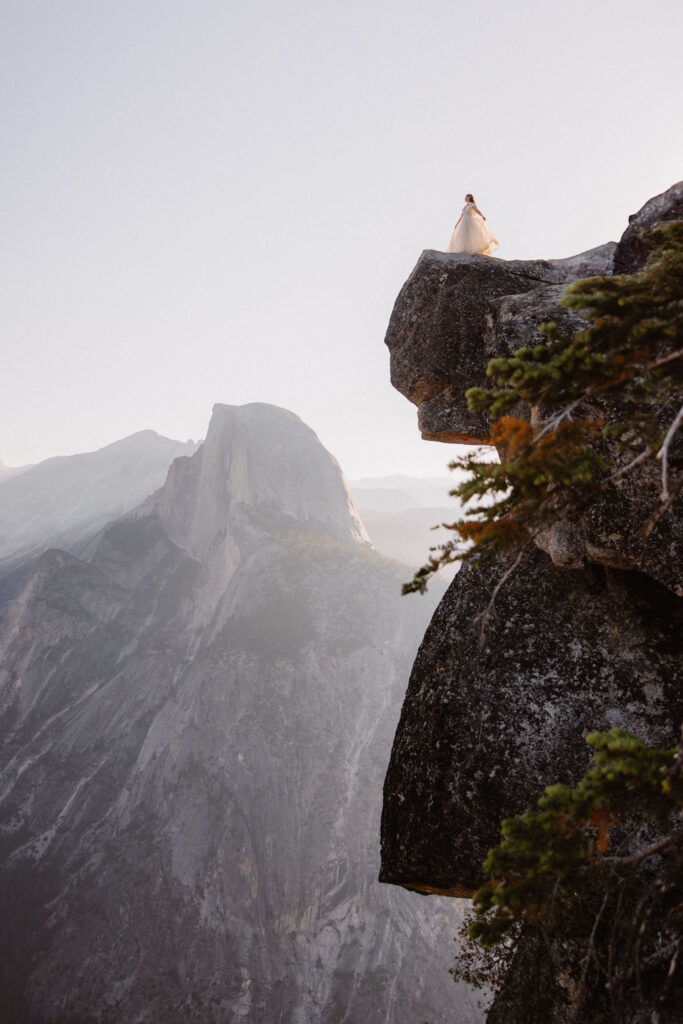 A couple in wedding attire stand together on a rocky overlook with a mountain landscape in the background at Yosemite national park at glacier point and mariposa grove