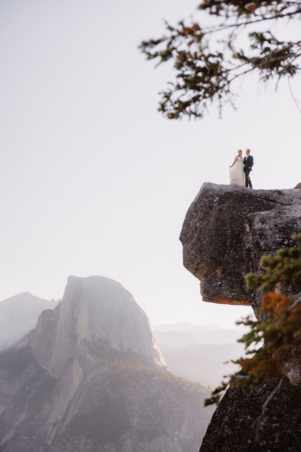 A couple in wedding attire stand together on a rocky overlook with a mountain landscape in the background at Yosemite national park at glacier point and mariposa grove