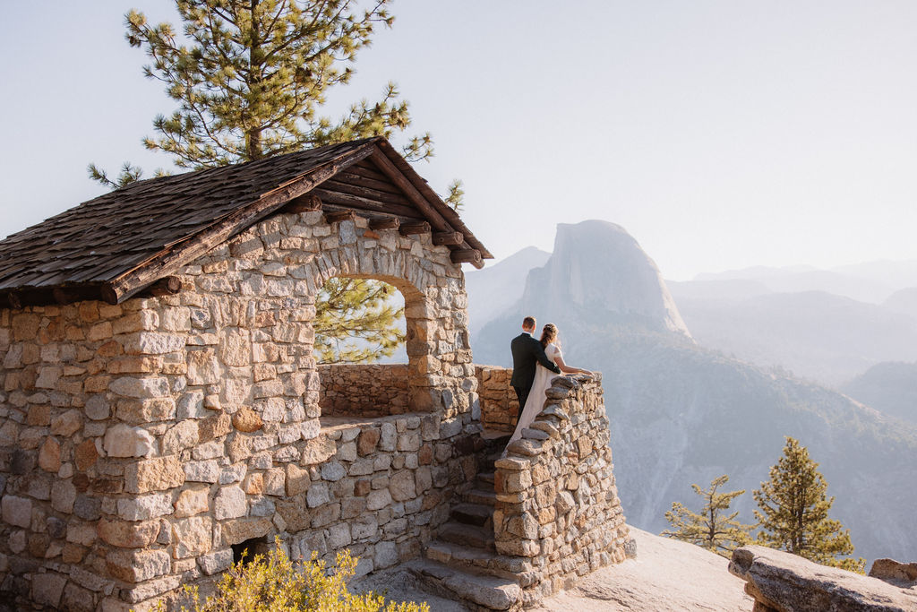 Two people sit on a stone building's ledge overlooking a mountainous landscape with trees and a clear sky.