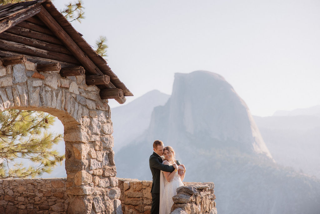 Two people sit on a stone building's ledge overlooking a mountainous landscape with trees and a clear sky.