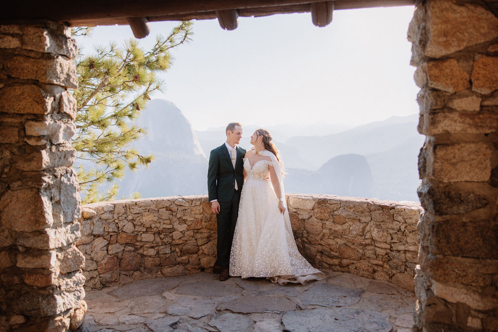 Two people sit on a stone building's ledge overlooking a mountainous landscape with trees and a clear sky.