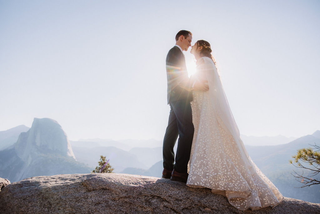 Two people sit on a stone building's ledge overlooking a mountainous landscape with trees and a clear sky.