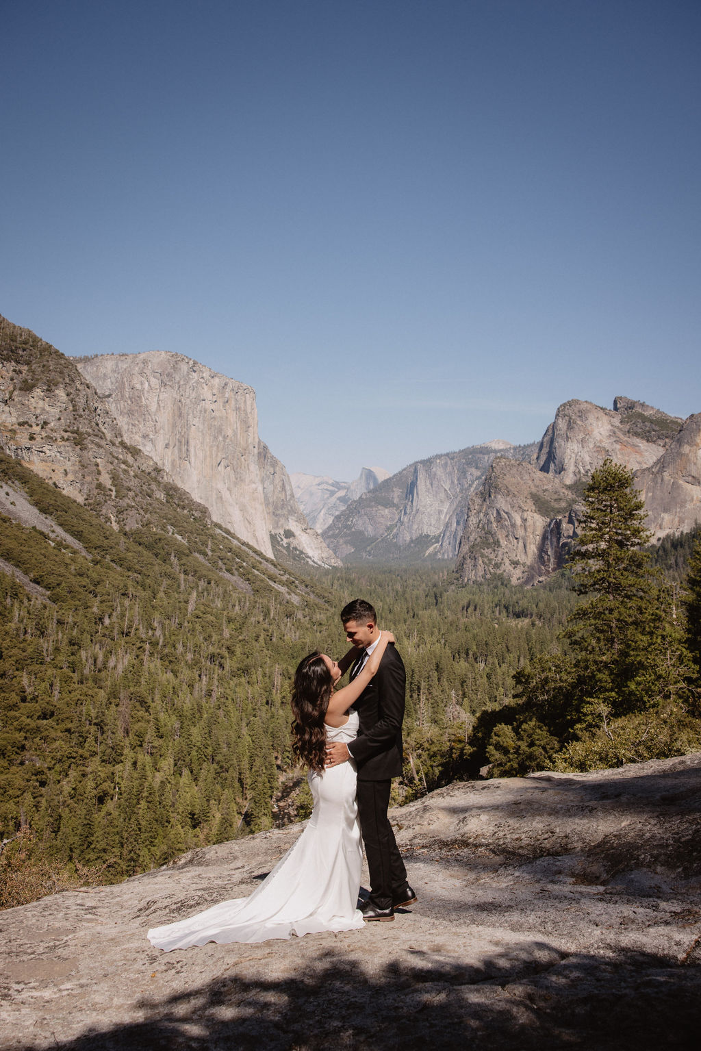 A couple dressed in wedding attire stands on a mountain ledge overlooking a vast rocky landscape with a prominent peak in the background. Best views in yosemite national park