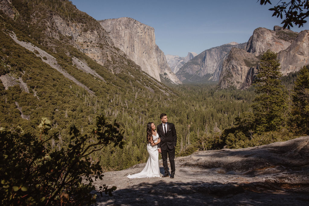 A couple dressed in wedding attire stands on a mountain ledge overlooking a vast rocky landscape with a prominent peak in the background. Best views in yosemite national park