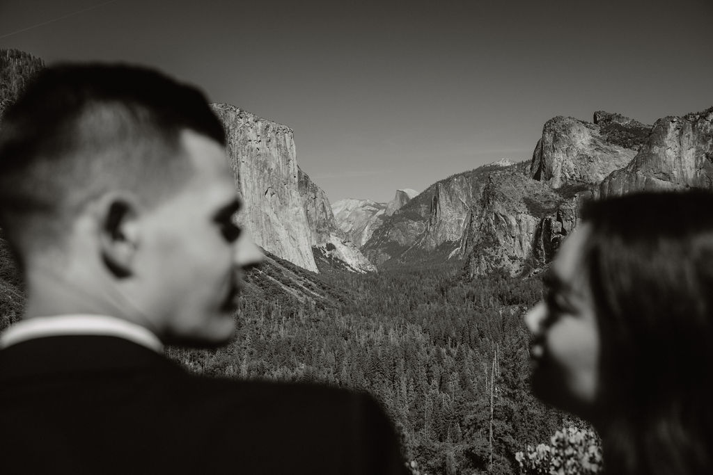 A couple dressed in wedding attire stands on a mountain ledge overlooking a vast rocky landscape with a prominent peak in the background. Best views in yosemite national park