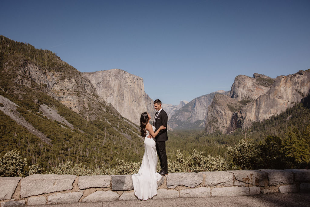 A couple dressed in wedding attire stands on a mountain ledge overlooking a vast rocky landscape with a prominent peak in the background. Best views in yosemite national park