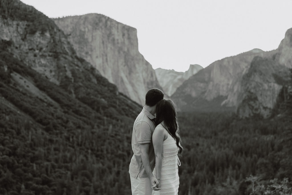 A couple stands embracing, facing towards a distant valley surrounded by large rocky mountains for their Yosemite National park engagement photos