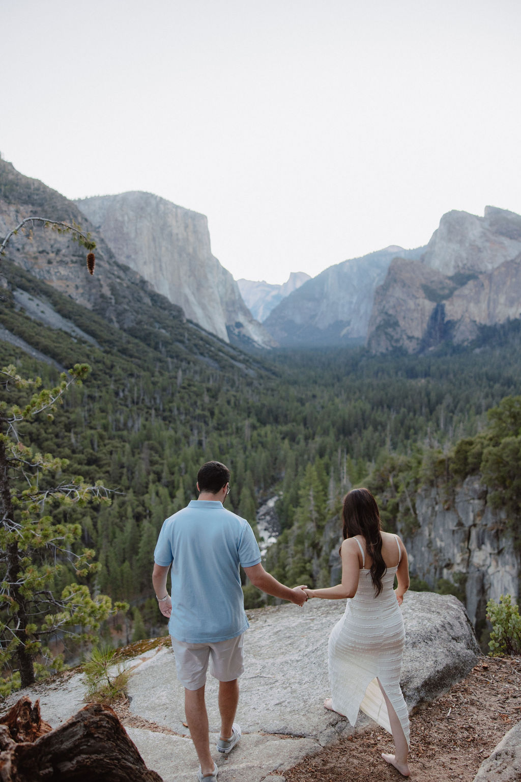 A couple holding hands stands on a rocky overlook, gazing at a valley with lush green forests and distant mountains for theor yosemite national park engagement photos