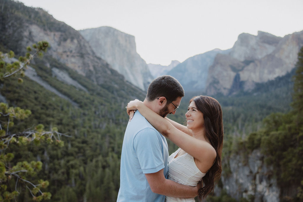 A couple smiling at each other in a mountainous landscape, with trees and cliffs in the background Yosemite National Park Engagement Photos 