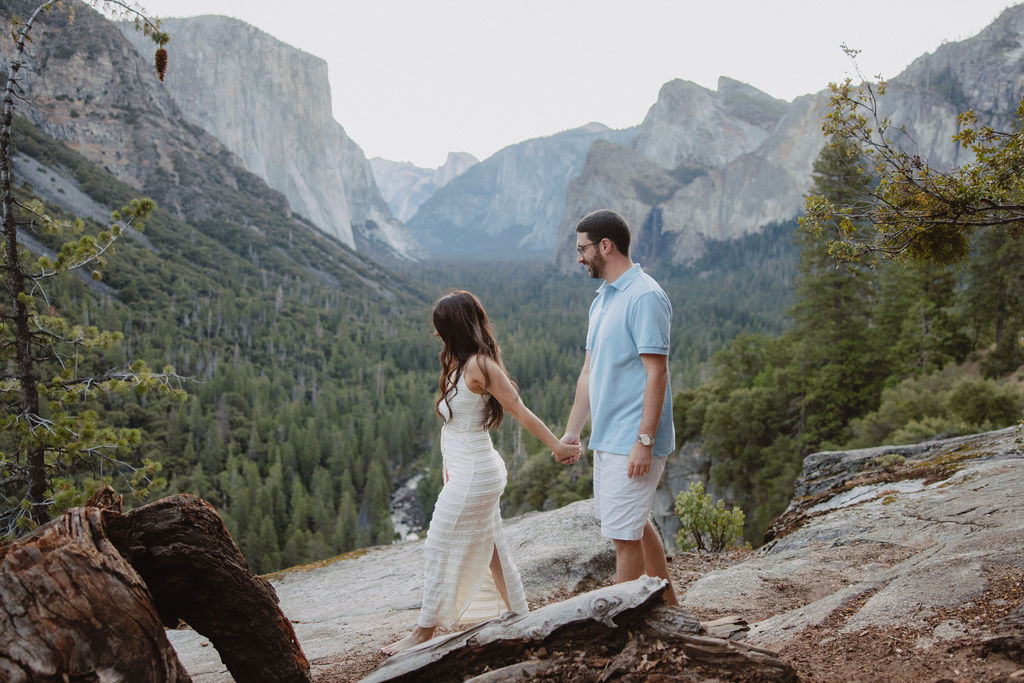 A couple holds hands while standing on a rocky ledge overlooking a forested valley and distant mountains for their Yosemite national park engagement photos