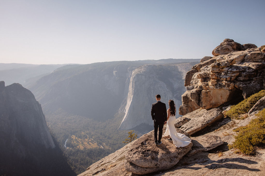 A couple dressed in wedding attire stands on a mountain ledge overlooking a vast rocky landscape with a prominent peak in the background. Best views in yosemite national park