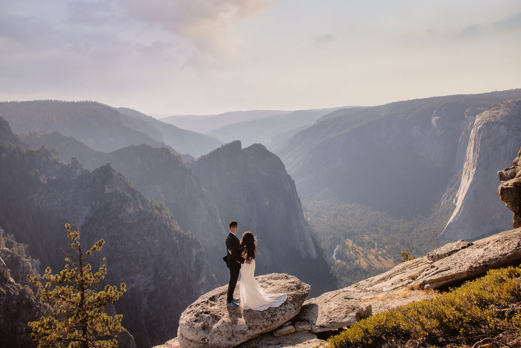 A couple dressed in wedding attire stands on a mountain ledge overlooking a vast rocky landscape with a prominent peak in the background. Best views in yosemite national park