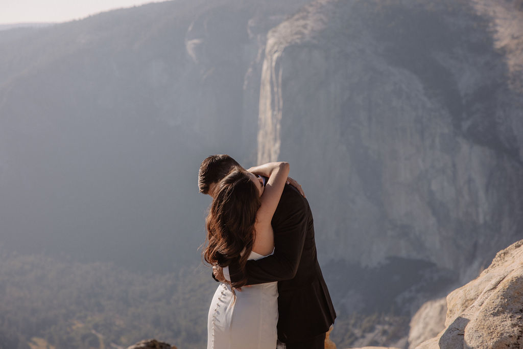 A couple dressed in wedding attire stands on a mountain ledge overlooking a vast rocky landscape with a prominent peak in the background. Best views in yosemite national park