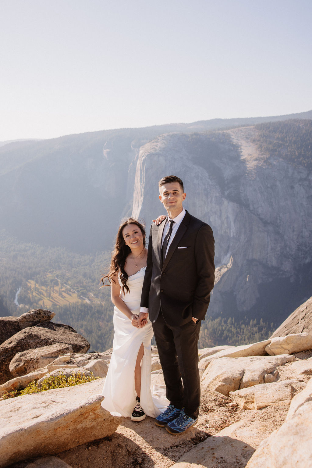 A couple dressed in wedding attire stands on a mountain ledge overlooking a vast rocky landscape with a prominent peak in the background. Best views in yosemite national park