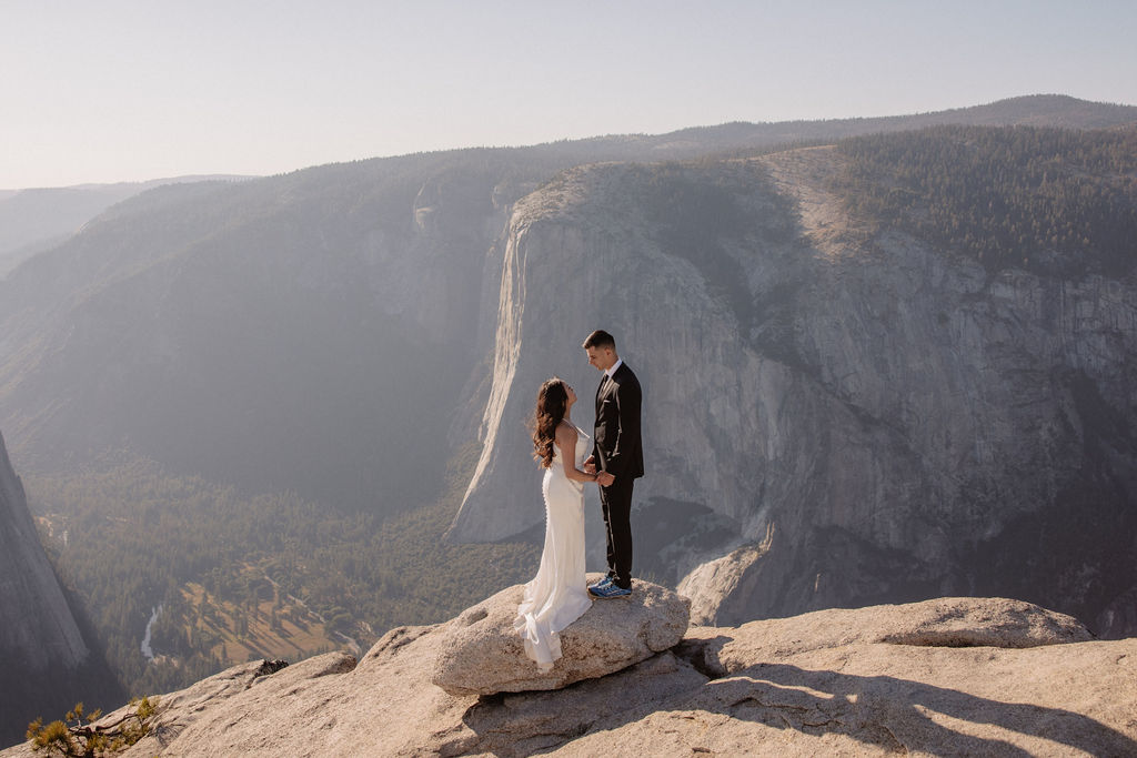 A couple dressed in wedding attire stands on a mountain ledge overlooking a vast rocky landscape with a prominent peak in the background. Best views in yosemite national park