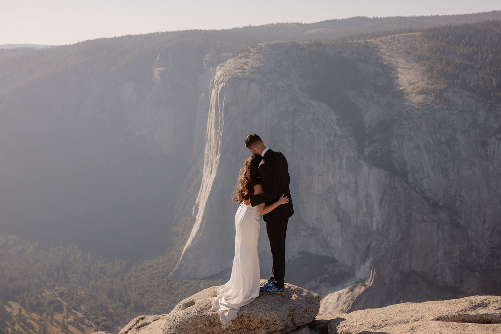 A couple dressed in wedding attire stands on a mountain ledge overlooking a vast rocky landscape with a prominent peak in the background. Best views in yosemite national park