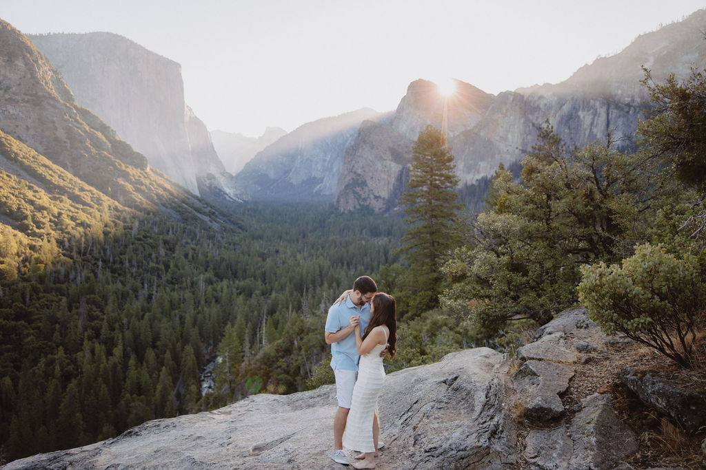 A couple embraces on a rocky ledge overlooking a valley with dense forests and mountains under a clear sky for their Yosemite national park engagement photos