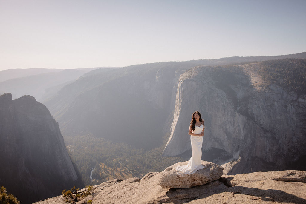 A couple dressed in wedding attire stands on a mountain ledge overlooking a vast rocky landscape with a prominent peak in the background. Best views in yosemite national park