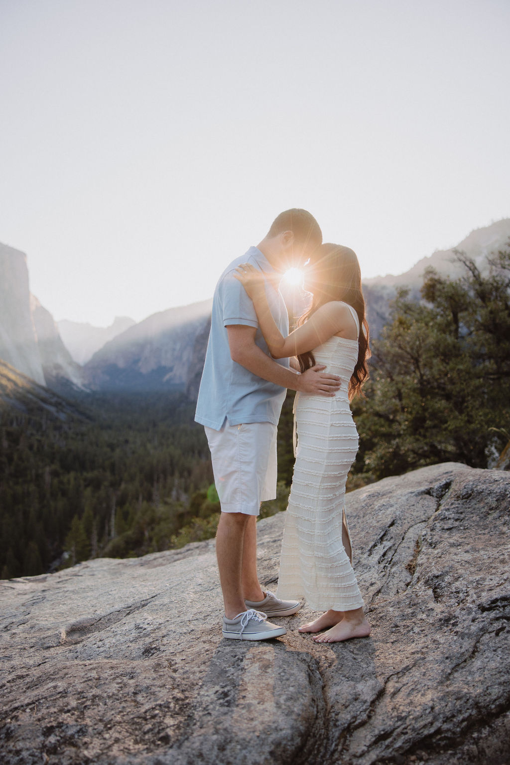 A couple stands on a rocky outcrop in a scenic mountainous area, embracing as the sun sets behind them.