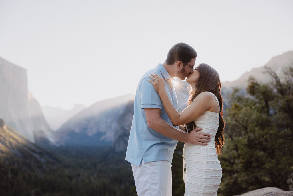 A couple kisses in front of a scenic mountain landscape under a clear sky, with trees and cliffs in the background for their Yosemite national park engagement photos