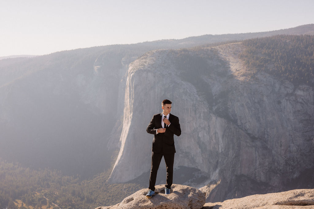 A couple dressed in wedding attire stands on a mountain ledge overlooking a vast rocky landscape with a prominent peak in the background. Best views in yosemite national park