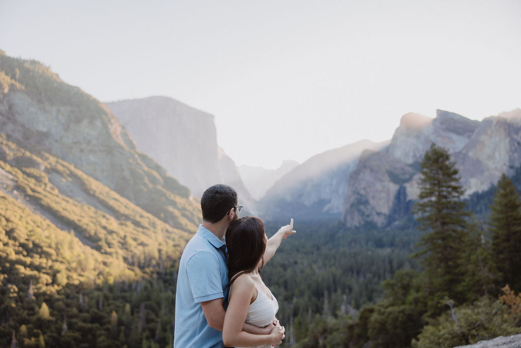 A couple kisses in front of a scenic mountain landscape under a clear sky, with trees and cliffs in the background for their Yosemite national park engagement photos